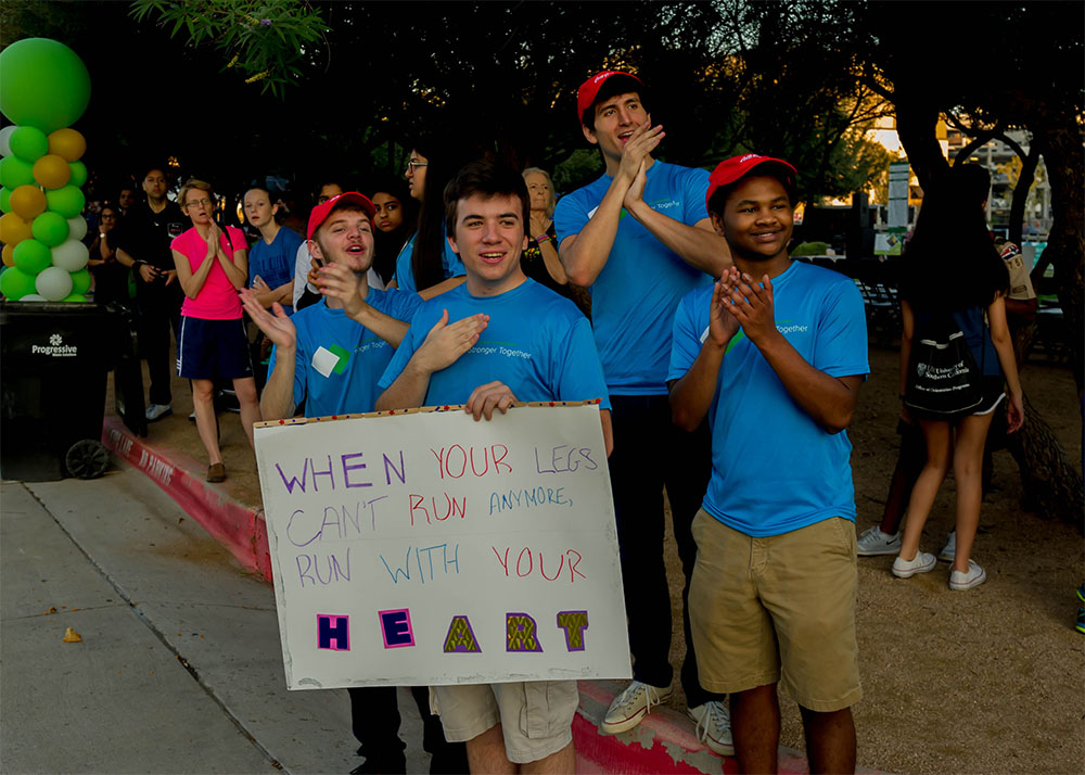 A group of young men clap for runners while holding a sign that reads 'When Your Legs Can't Run Anymore, Run with your HEART.'