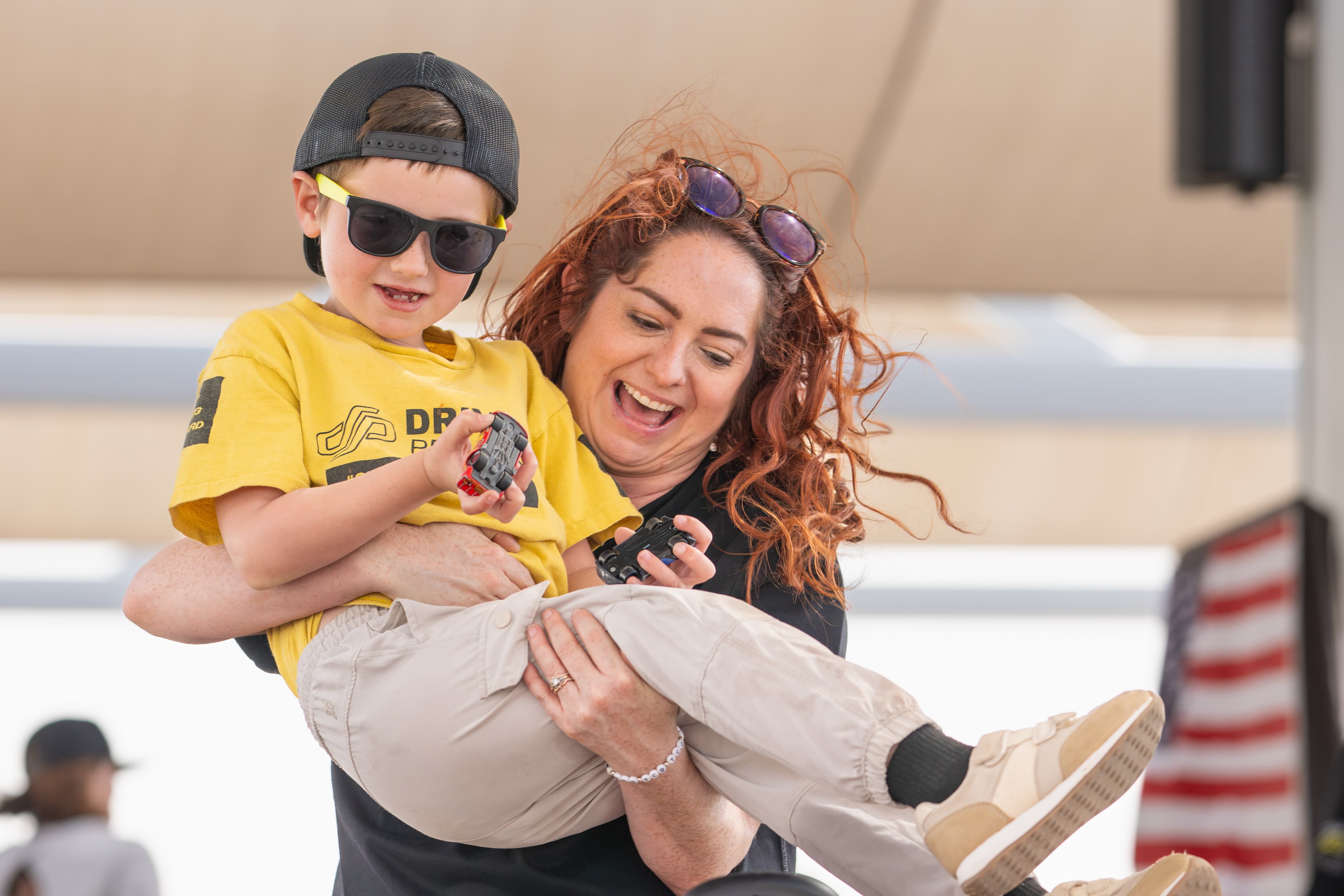 An adult carrying a child in a yellow Driven Project shirt holding a toy car. Both are smiling.