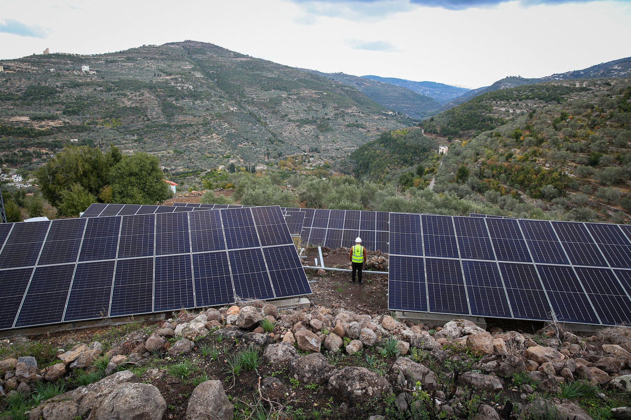 An array of solar panels in the hills in Syria. A worker wearing an Aga Khan Foundation hi-viz vest stands at center.
