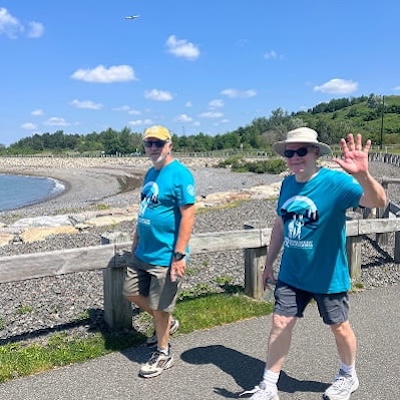 Two people in teal event shirts walk along a waterfront path on a sunny day, one waving at the camera.
