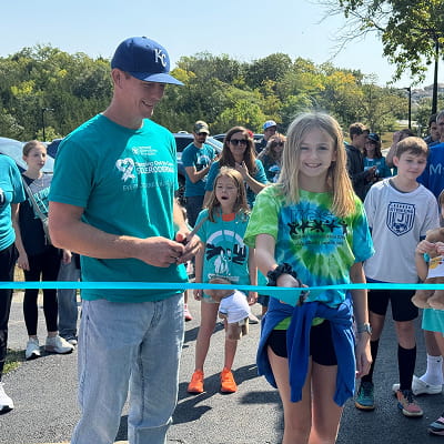 Man and girl cut a teal ribbon at an outdoor event surrounded by participants.