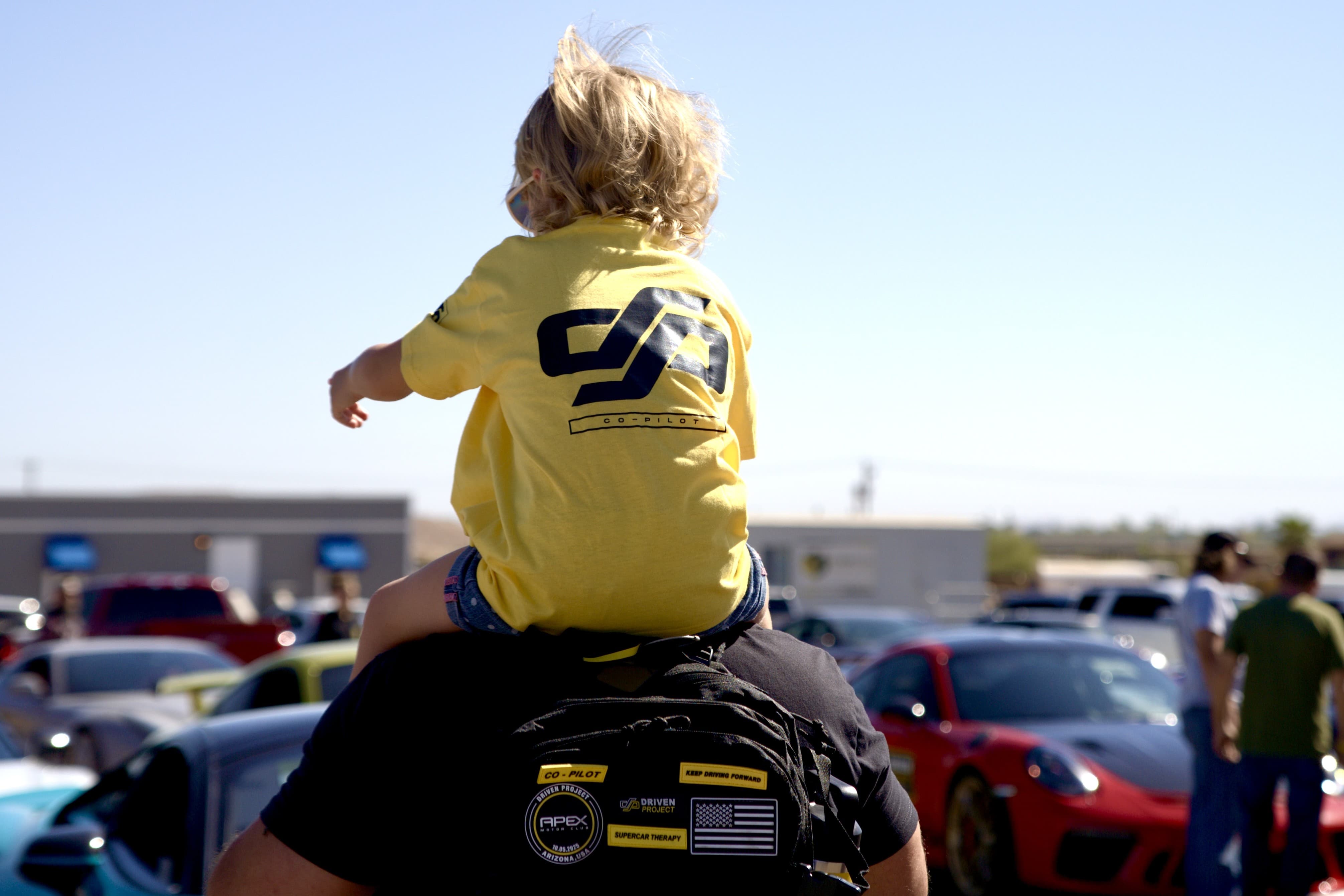 Child in a yellow Driven Project shirt riding on the shoulders of an adult. Both are facing away from the camera.