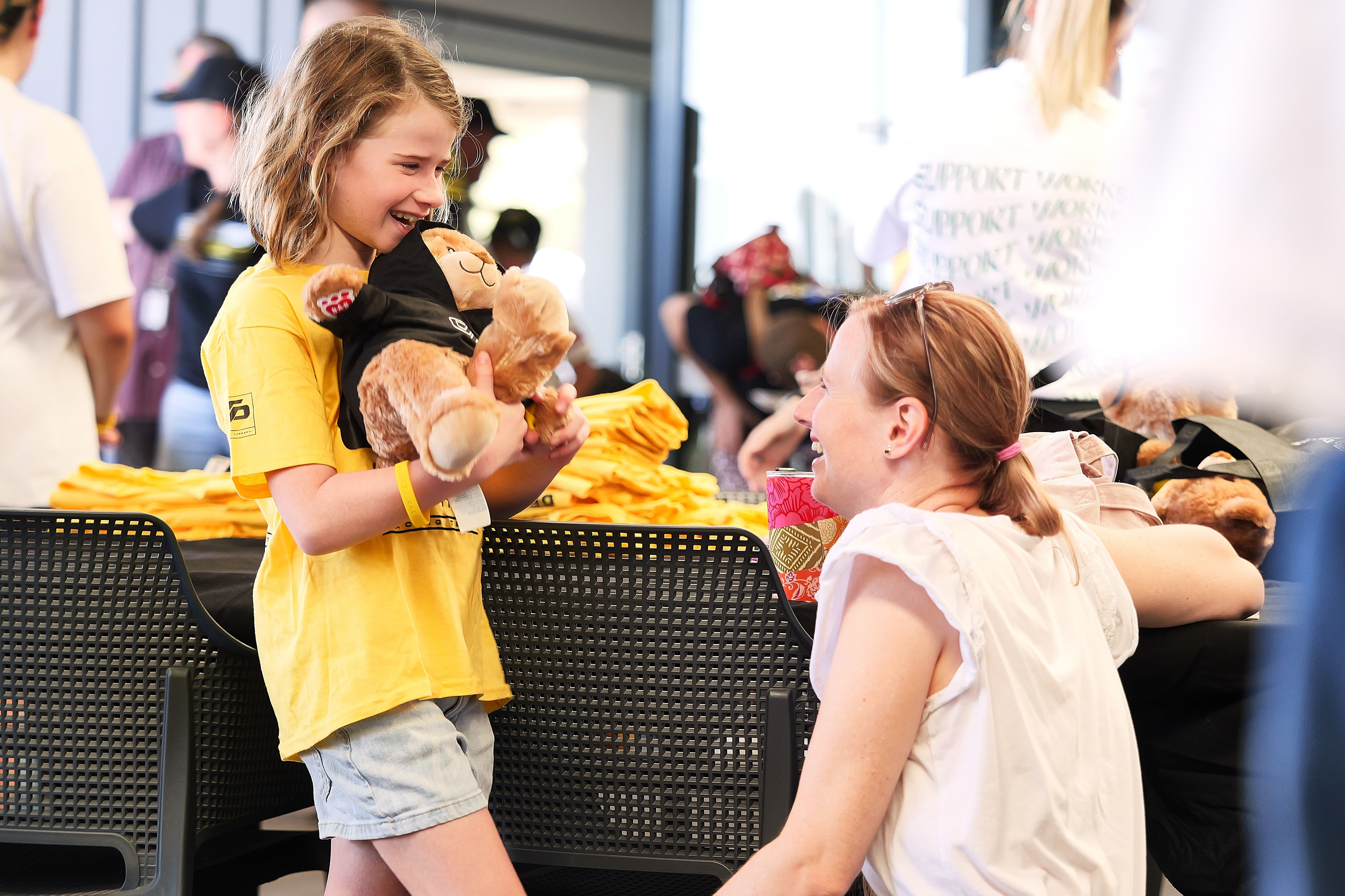 An child in a yellow Driven Project shirt holding a Driven Project teddy bear smiling at her parent.