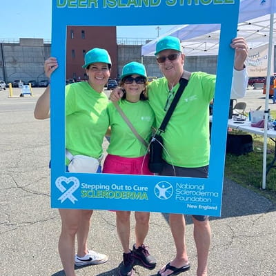 Three people in bright green shirts and teal hats pose together holding a “Deer Island Stroll” event frame.