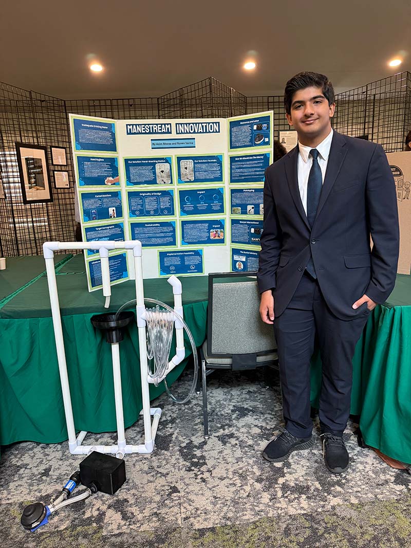 A young man stands next to a presentation board and prototype of an innovation A young man stands next to a presentation board and prototype of an innovation